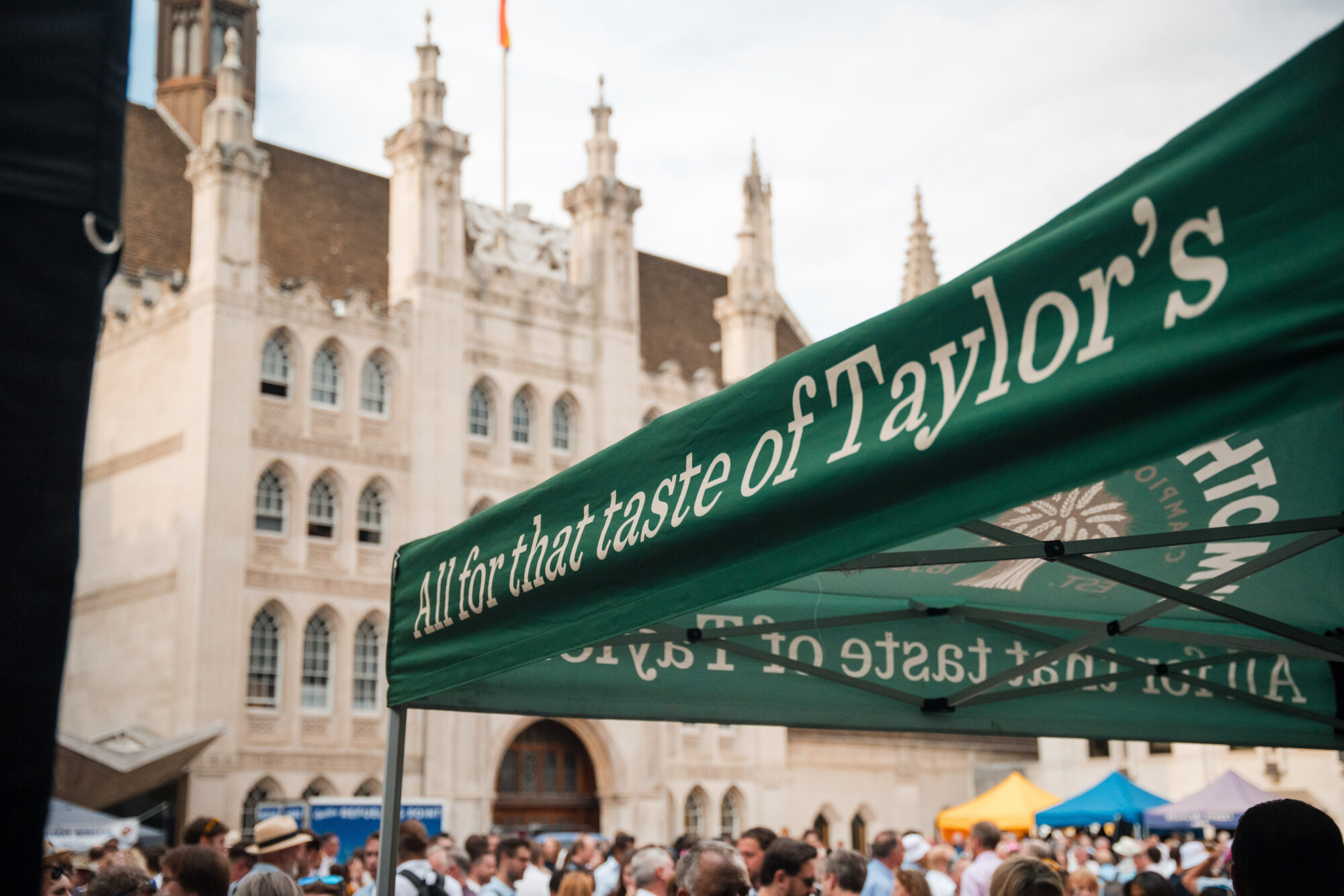 Outdoor event with a crowd in front of a historic building. A green canopy displays the words 