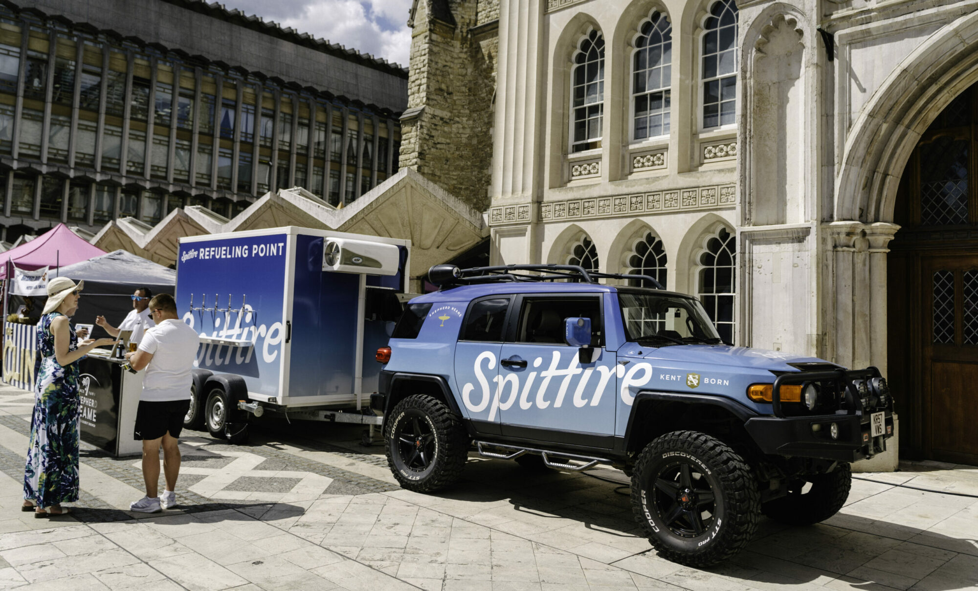 A branded blue car with a trailer is parked in a historic courtyard, with a group of people assembled behind the vehicle.
