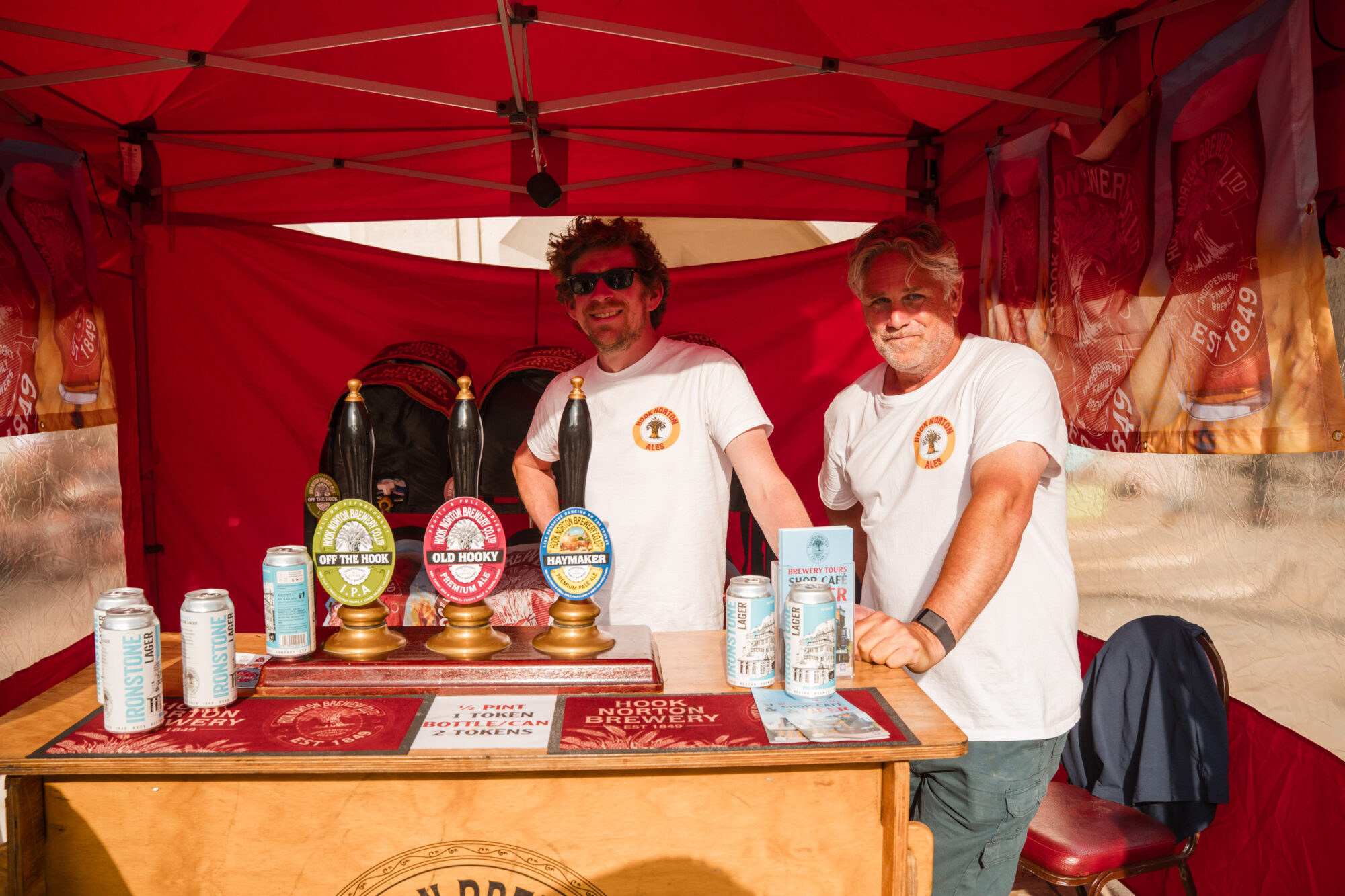 A brewery festival stall featuring a selection of beer taps and cans from Hook Norton Brewery. There are two people standing behind the counter under a red canopy.