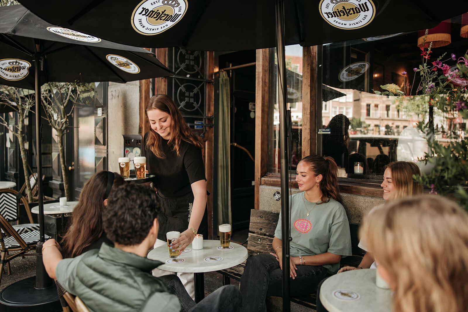A person serves drinks to a group sitting at a cafe table outdoors. Several glasses of beer are on the table.