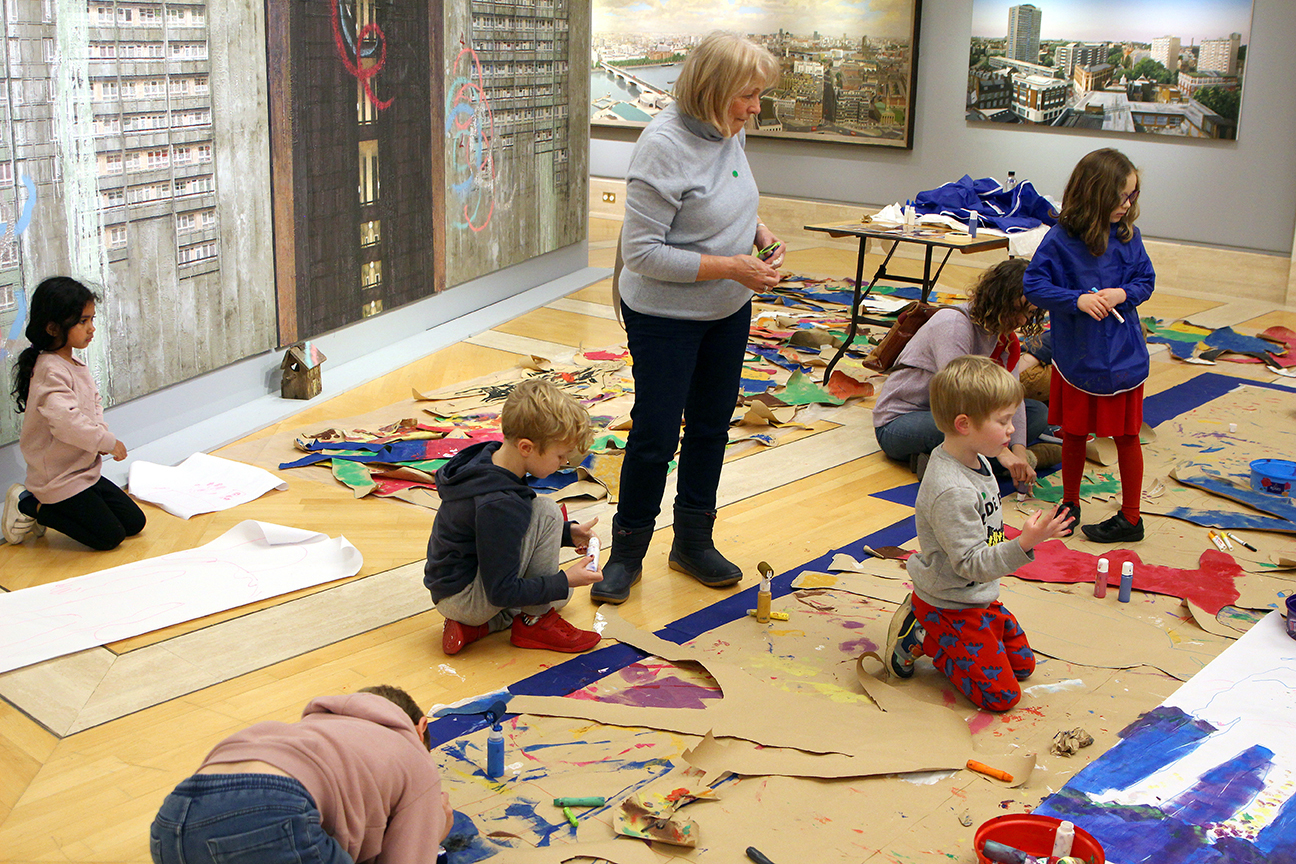 Children and an adult engaged in art activities on a gallery floor filled with colorful paper cutouts. Large framed artworks of urban scenes are displayed on the wall.