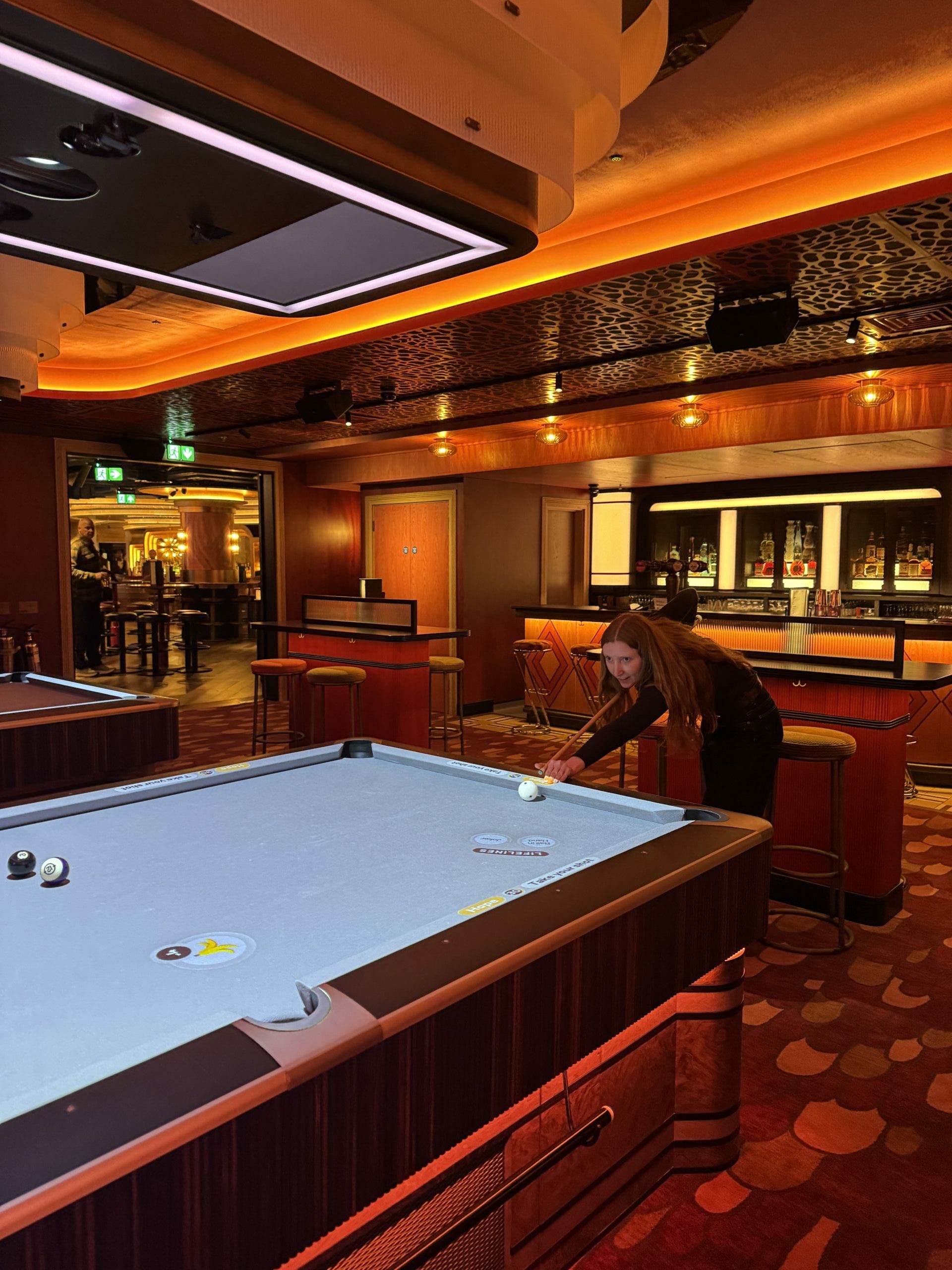 someone holding a pool cue aiming it at a ball on a pool table in a grand setting with a bar in the background