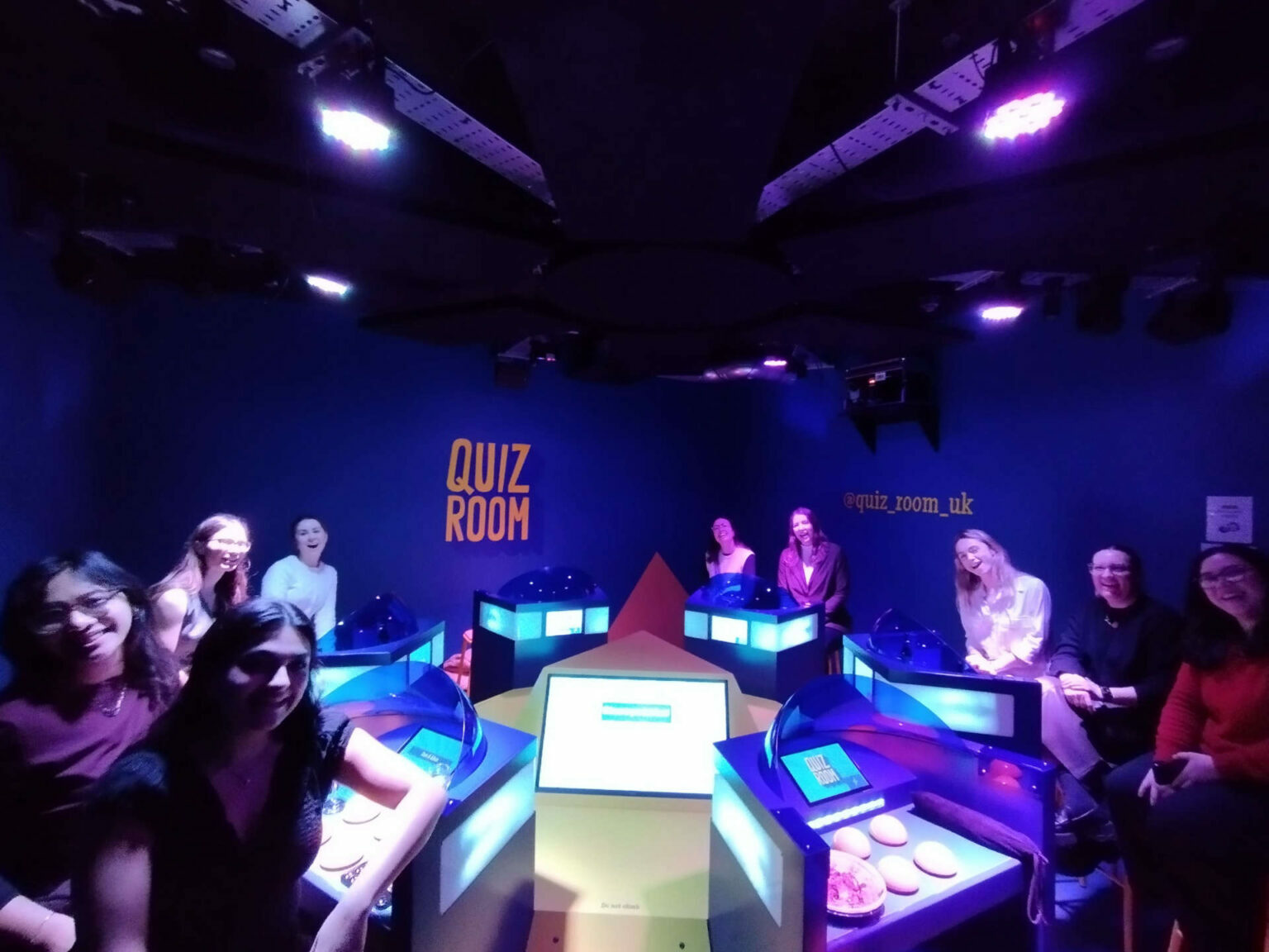 An image in a coloured lit up room with teams sat around a central console smiling for the cameras